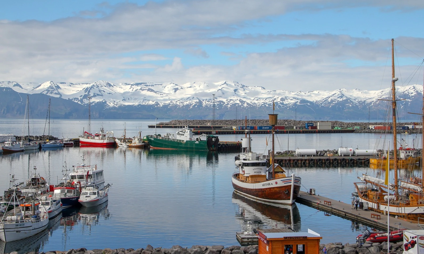 A picturesque harbour in Iceland with fishing boats docked on calm waters, surrounded by snow-capped mountains in the background.
