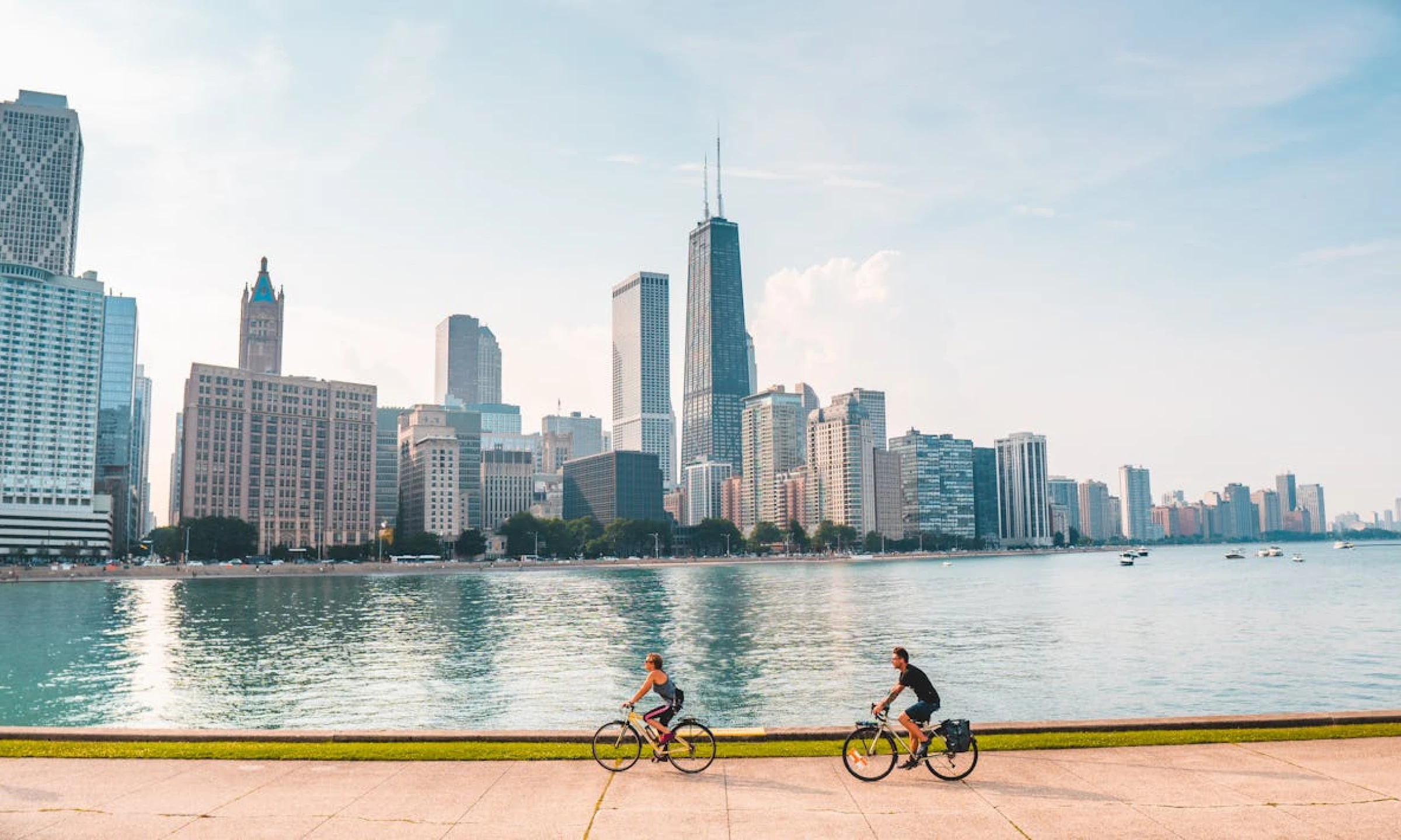 Two cyclists ride along a waterfront path with the Chicago skyline in the background. The water reflects the iconic skyscrapers, including the John Hancock Center, under a clear, bright sky. Boats are visible on the water.