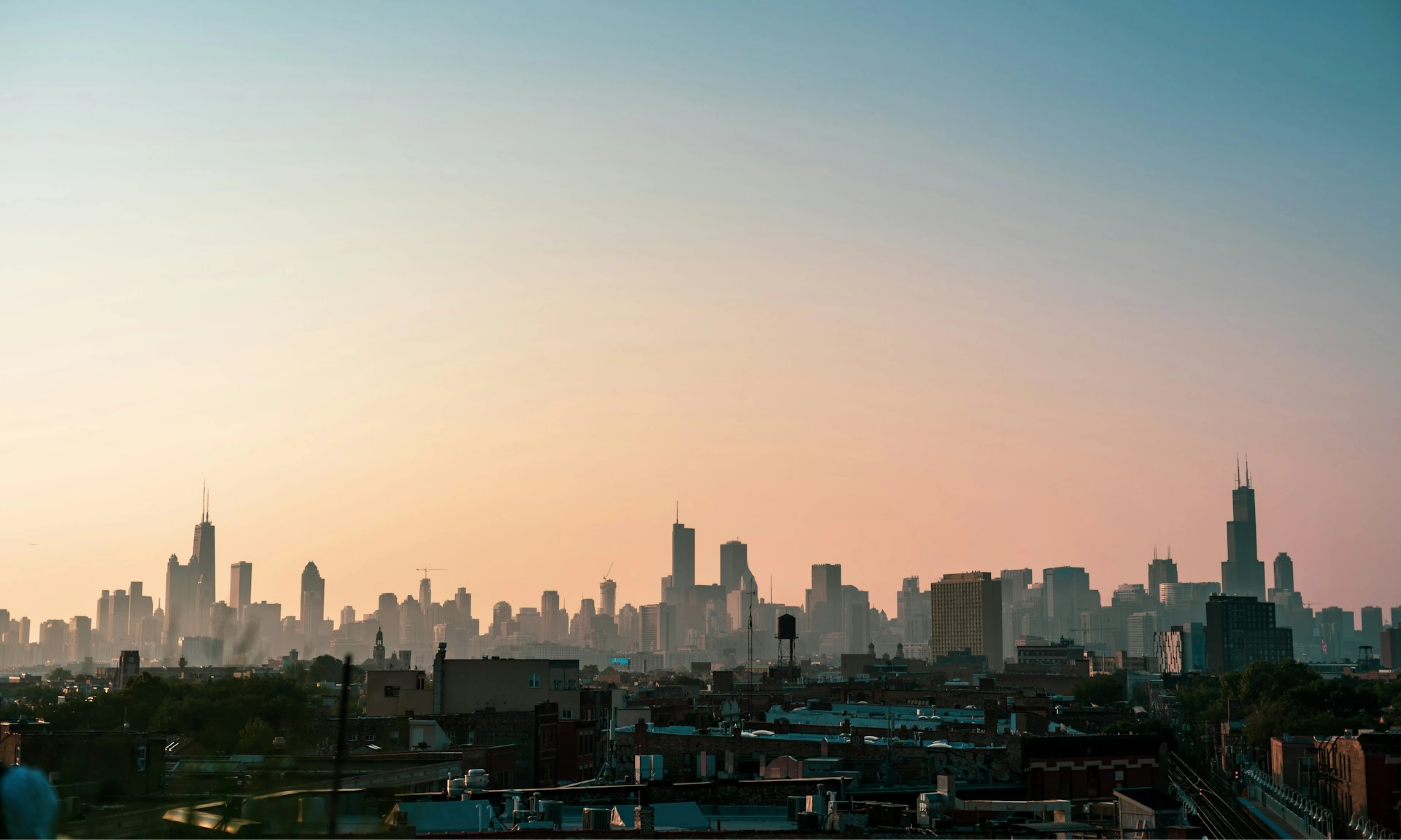 A panoramic view of the Chicago skyline at sunset, with the sky transitioning from warm oranges to cool blues. The city's distinctive skyscrapers, including the Willis Tower, stand prominently against the horizon.