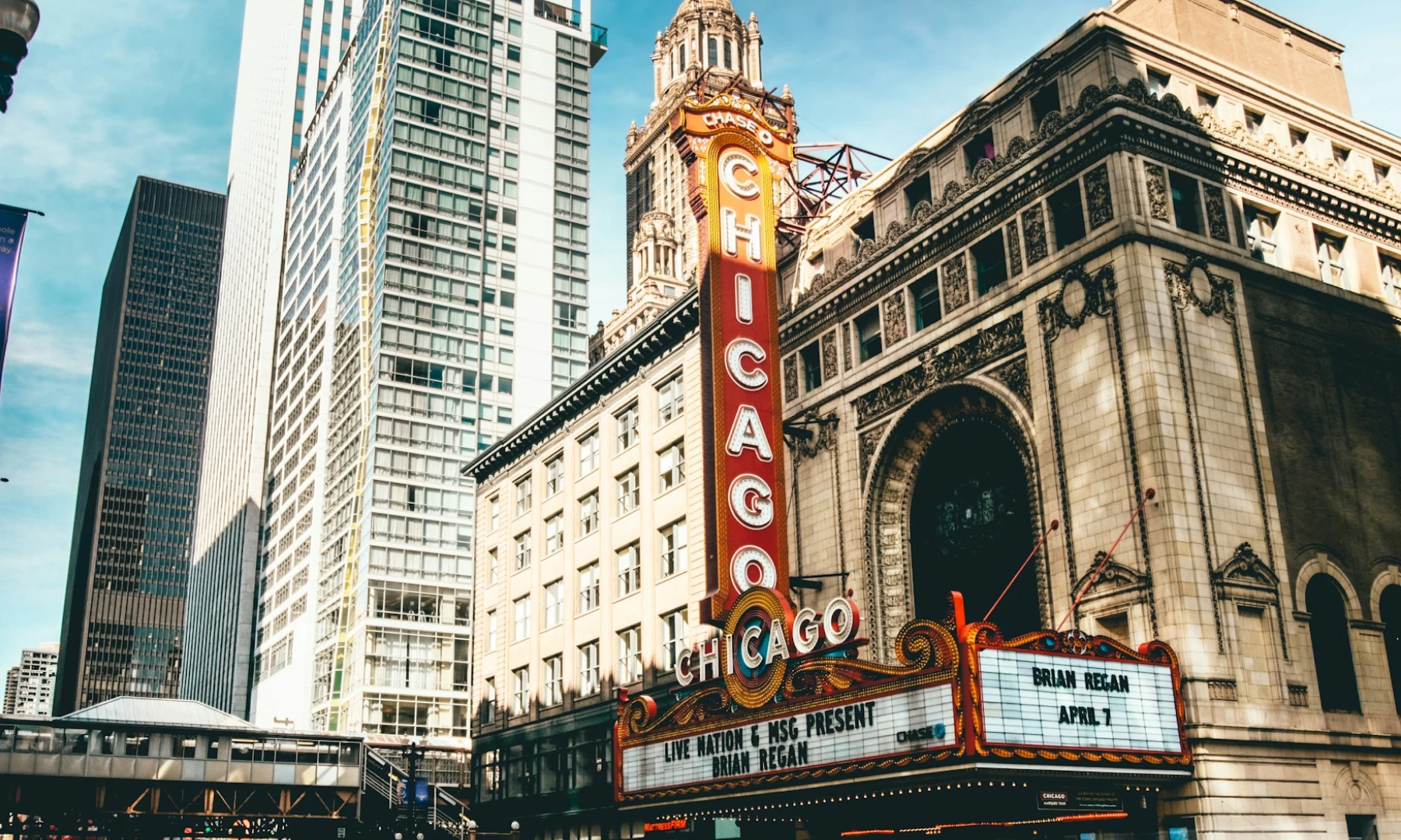 The iconic Chicago Theatre sign illuminated on a busy city street, with surrounding tall buildings reflecting the city's nightlife. This is one of the top places to visit on a Chicago overnight layover. The marquee advertises a performance by Brian Regan on April 1st. Traffic blurs by, highlighting the city's energetic pace.