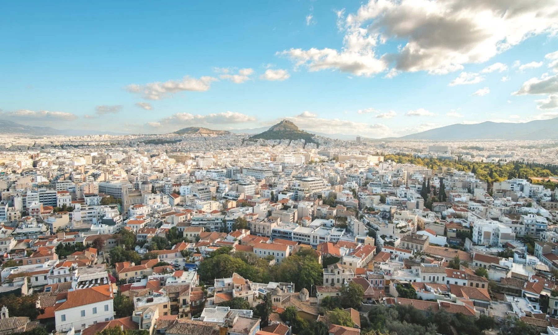 Aerial image of Athens, displaying a sprawling city with numerous buildings and hills under a partly cloudy sky.