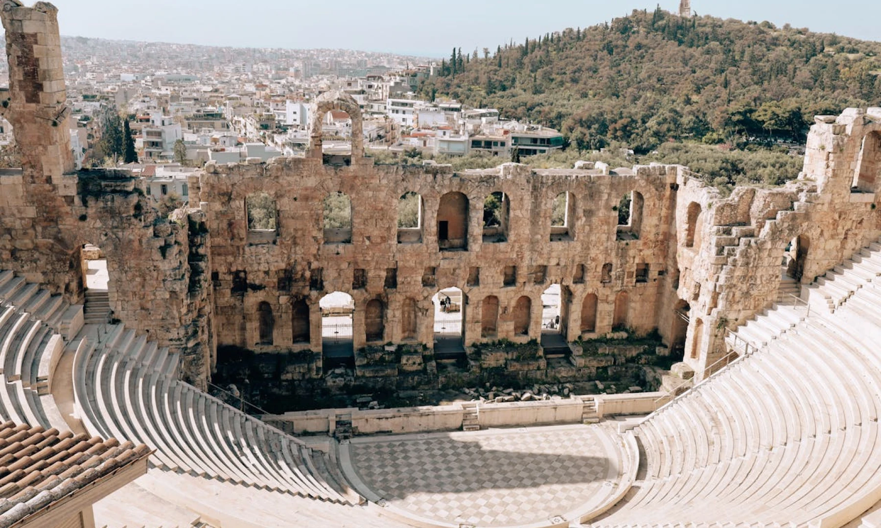 Image of the ancient Odeon of Herodes Atticus in Athens, with its semicircular seating arrangement and partially intact stone walls, with the modern cityscape and a forested hill in the background. This is one of the top sights on an Athens layover.