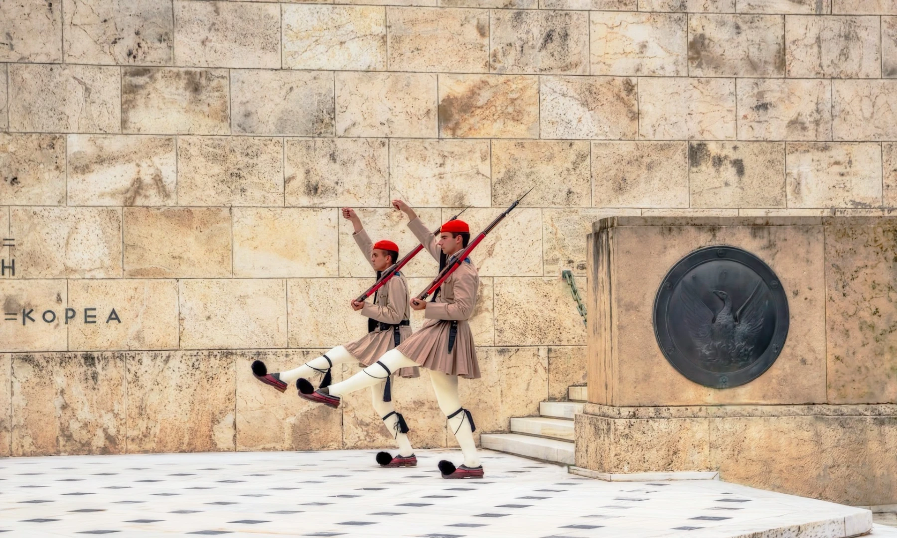 Image of two Greek Evzones soldiers performing the ceremonial guard change at the Tomb of the Unknown Soldier in Athens, wearing traditional uniforms with red berets, beige tunics, white kilts, and black pom-pom shoes, against a stone wall backdrop.