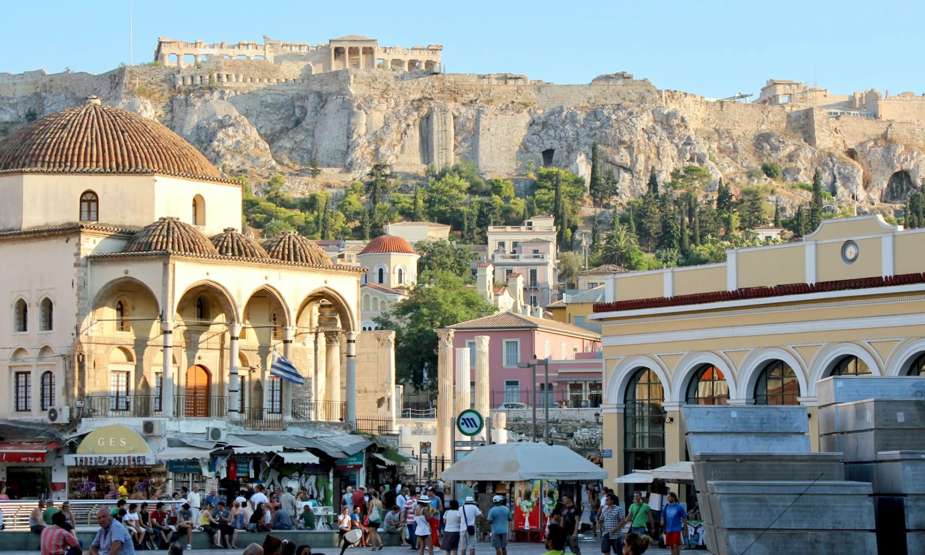 Image of a bustling city square in Athens with the Acropolis in the background, featuring a historical building with a domed roof and arches, surrounded by shops and people.