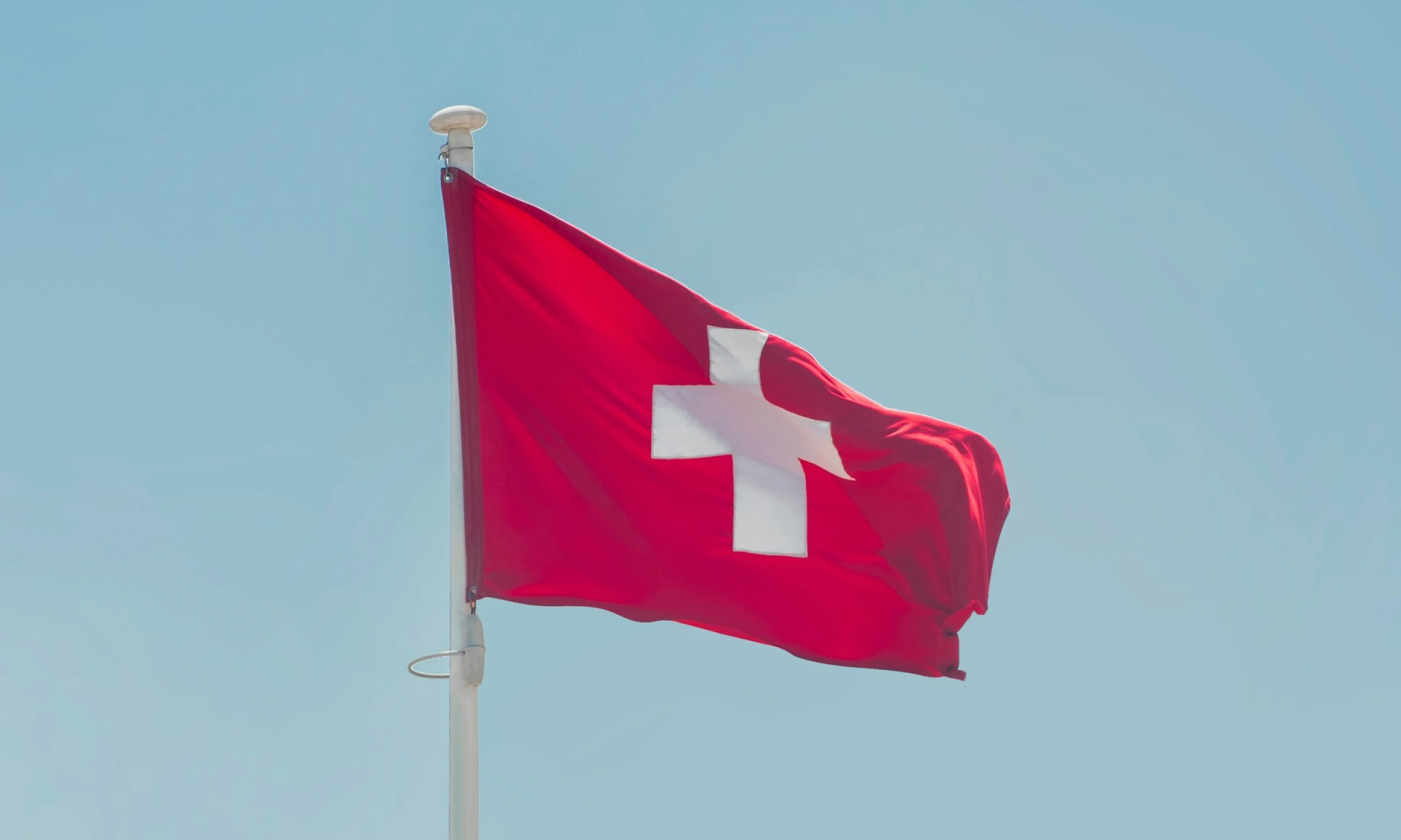 A Swiss flag fluttering against a clear blue sky with soft white clouds.
