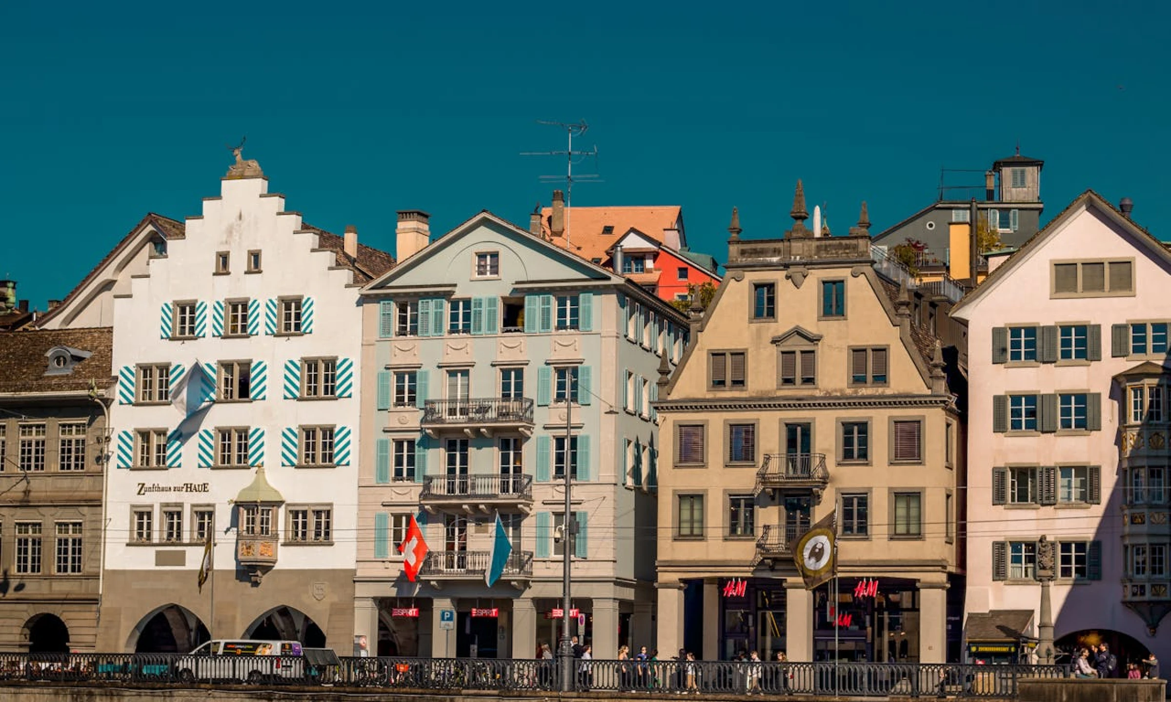 Historical buildings along a river in Zurich, with colourful facades and flags. The architecture includes white, pastel blue, and beige tones. This is one of the best areas to explore on a Zurich layover.