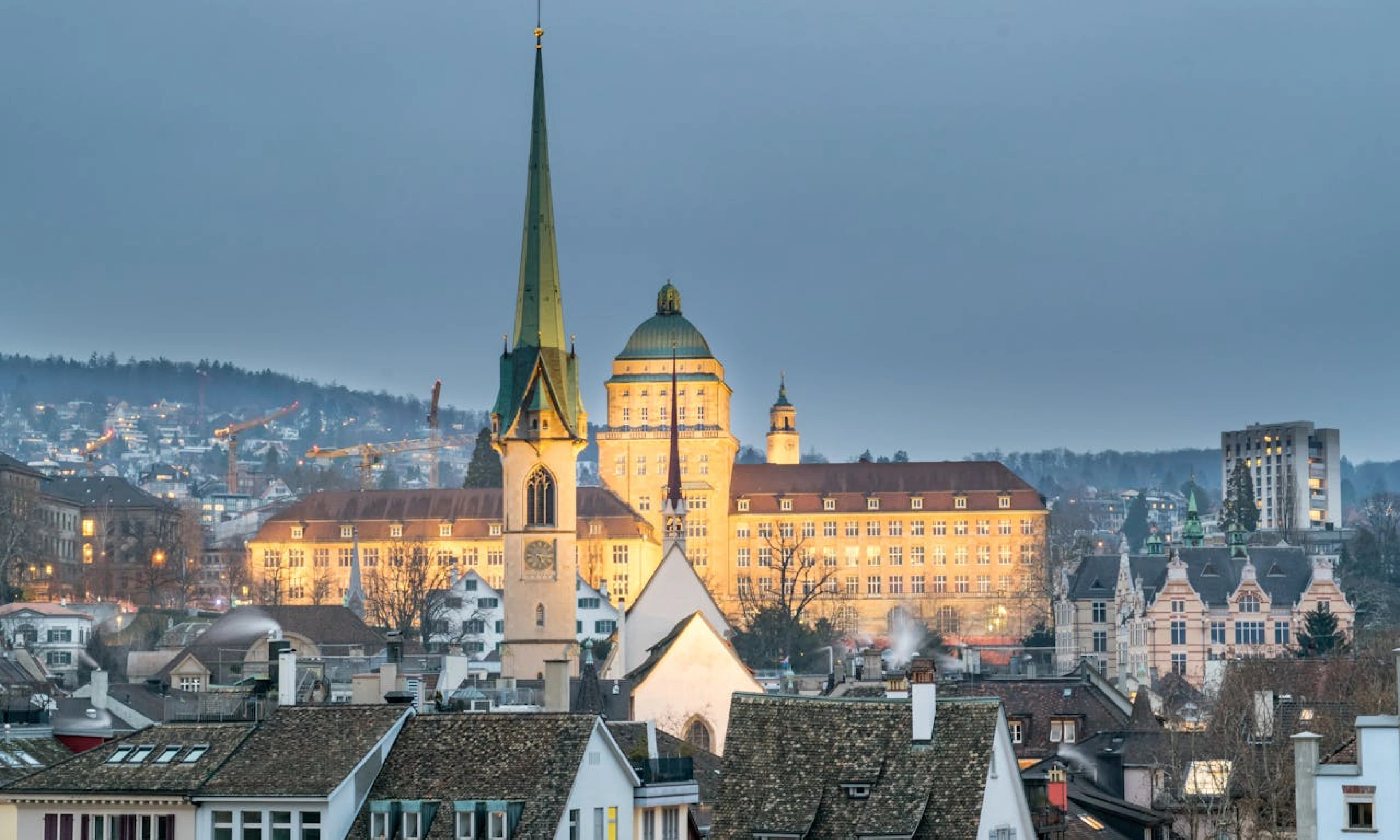 A panoramic view of Zurich at dusk, as seen on an overnight Zurich layover, featuring prominent buildings such as a church with a green spire and a large dome-shaped structure in the background. The city is lit with a soft glow against a dimming sky.