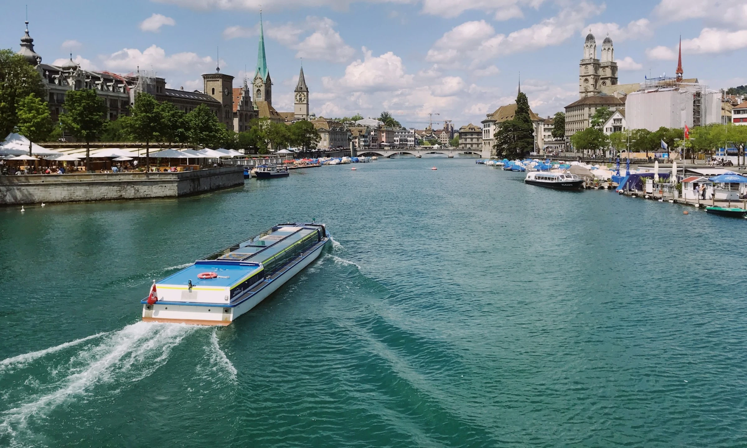 View of Zurich's waterfront featuring a boat cruising on the turquoise waters of the river. The cityscape includes historic buildings and a riverside promenade under a clear blue sky.
