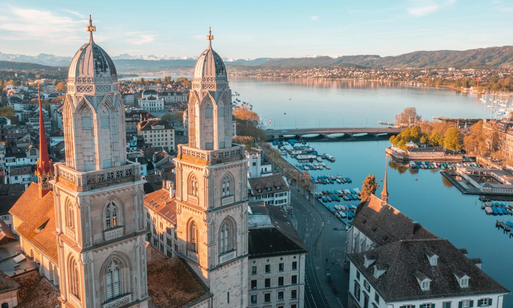 Aerial view of the Grossmünster Cathedral in Zurich during sunset, showcasing its twin towers with silver-coloured roofs and gold accents. Below, the city stretches towards Lake Zurich, lined with trees and buildings. This is one of the top places to visit on a Zurich layover.