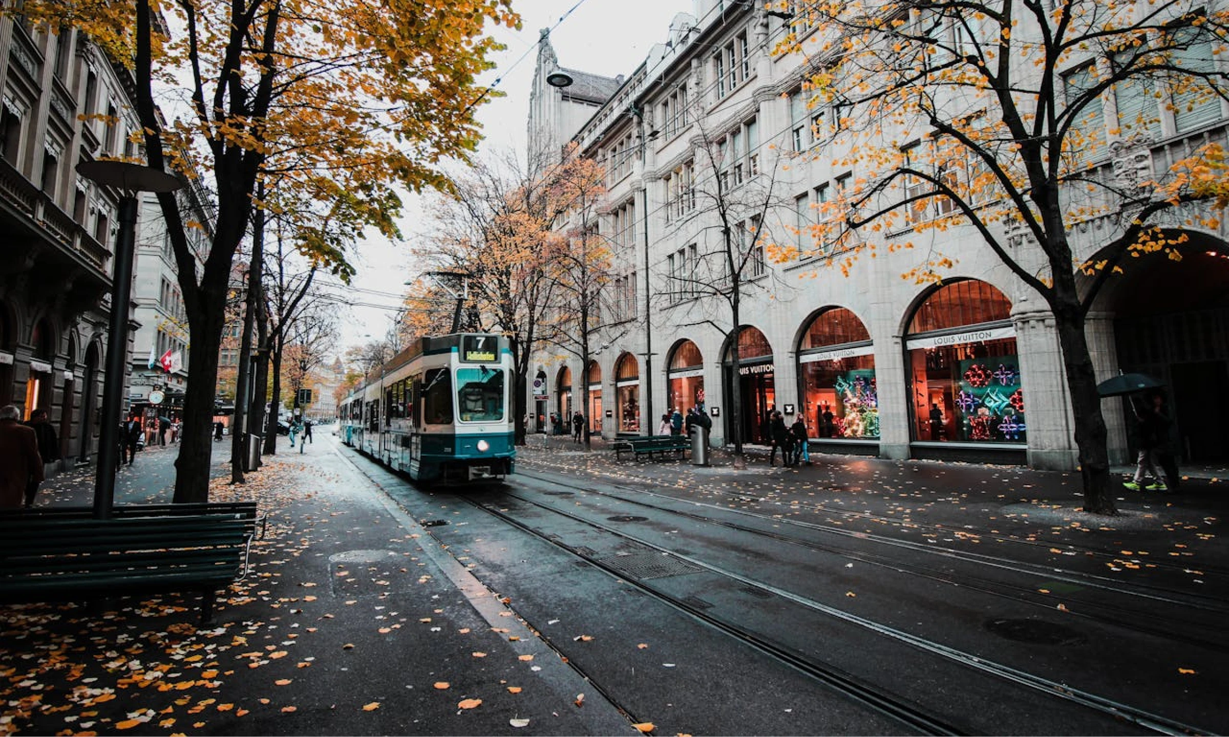 A tram travels down a tree-lined street in Zurich during autumn, with fallen yellow leaves covering the tracks and road. Buildings with shops line the pavements under a cloudy sky.