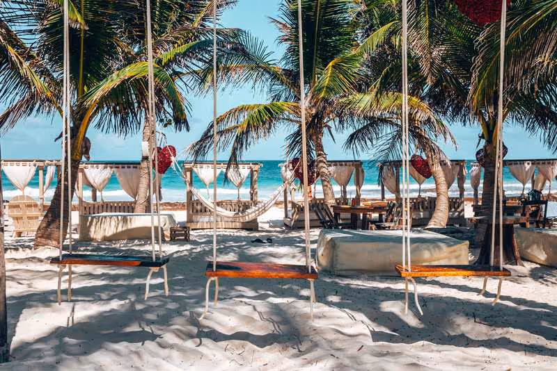 Plage de sable blanc à Tulum avec balançoires, palmiers, lits balinais et cabanes en toile face à la mer turquoise.