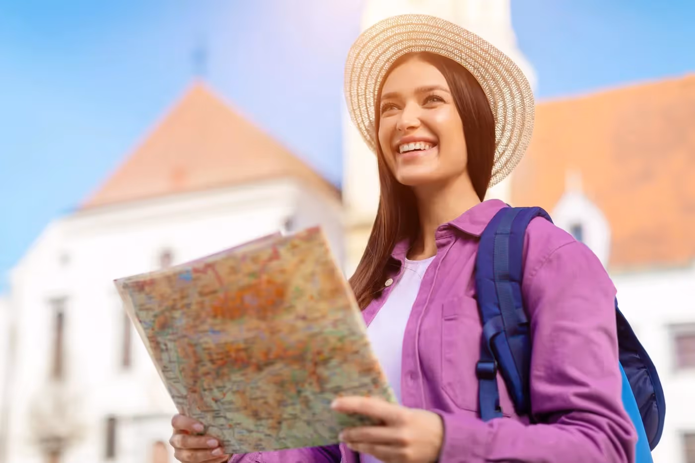 Jeune voyageuse souriante lisant une carte touristique en ville, portant un sac à dos et un chapeau de paille, devant des bâtiments historiques.