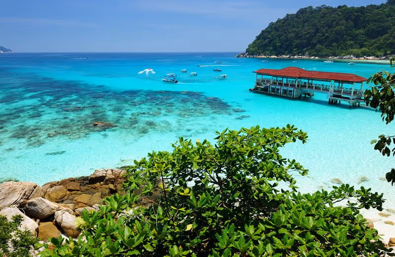 Eau turquoise cristalline bordée de verdure tropicale, bateaux de plaisance et ponton en bois sur une plage en Malaisie.