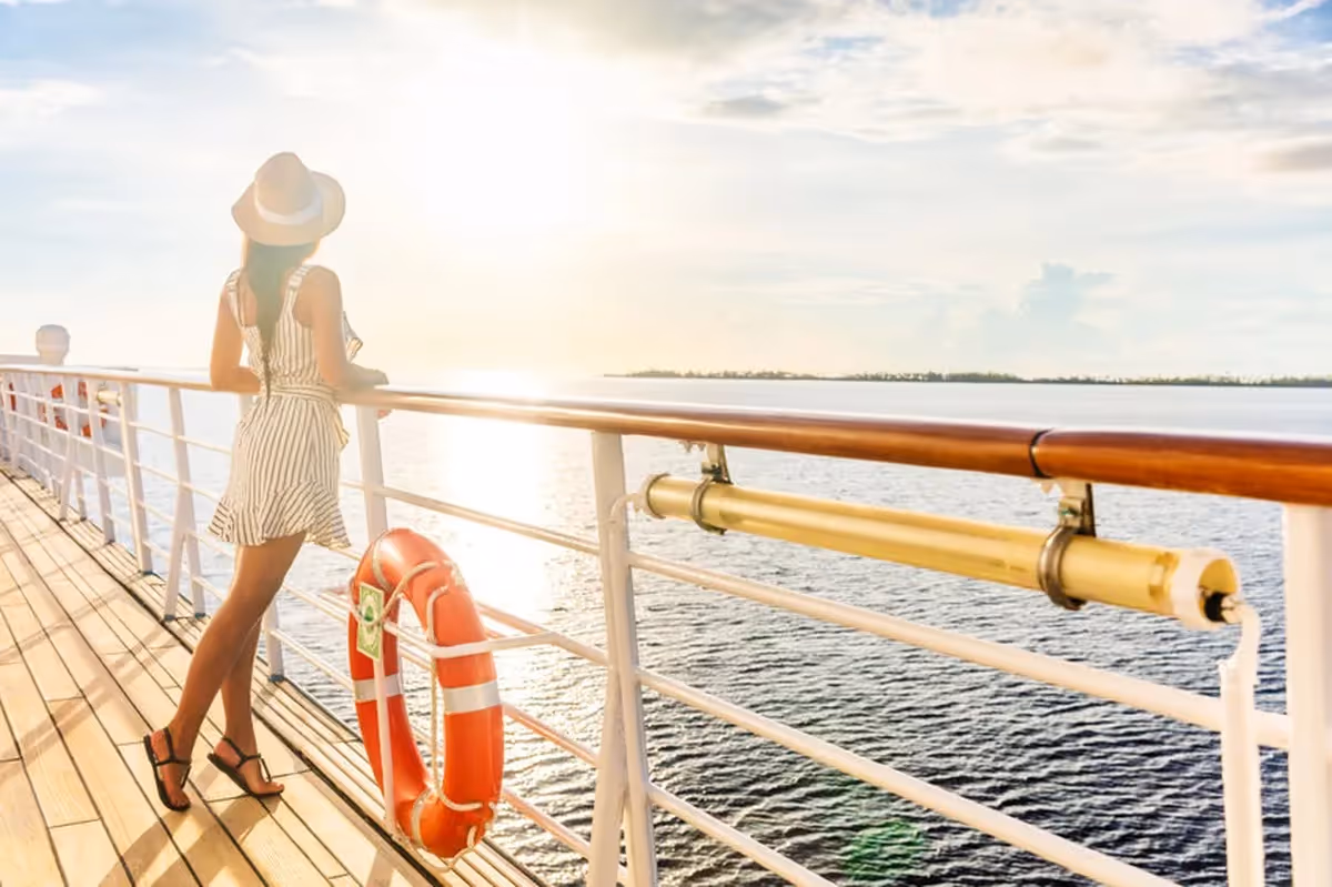 Femme en robe d’été et chapeau regardant l’horizon depuis le pont d’un bateau de croisière au coucher du soleil.
