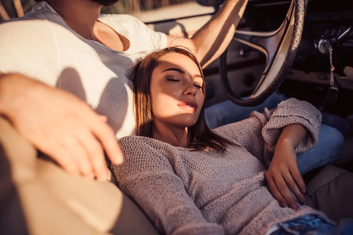 Jeune femme reposée, allongée contre son partenaire dans une voiture, symbolisant un moment paisible et complice pendant un road trip en amoureux.