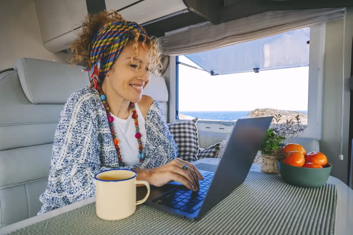 Femme nomade numérique travaillant sur un ordinateur portable dans un van face à la mer, tasse de café et fruits frais à ses côtés.