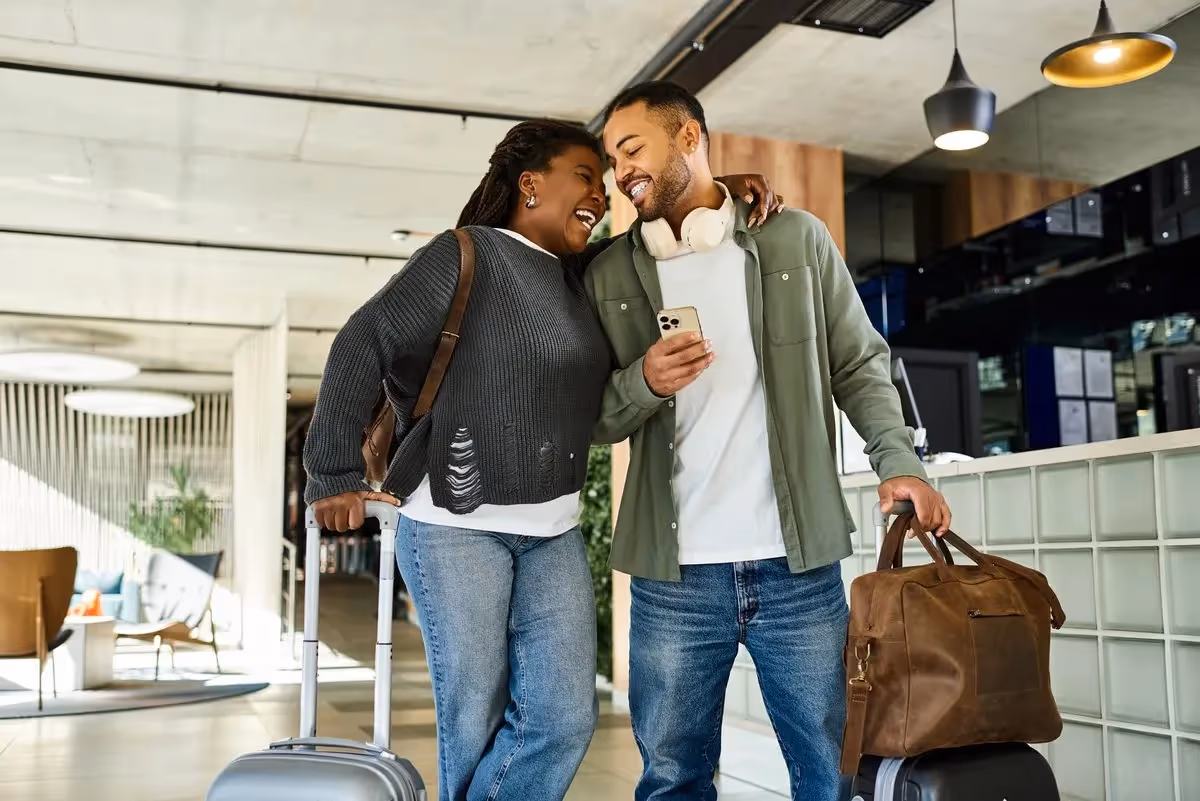 Un couple souriant arrive à l’hôtel avec ses valises, prêt pour un voyage de dernière minute.