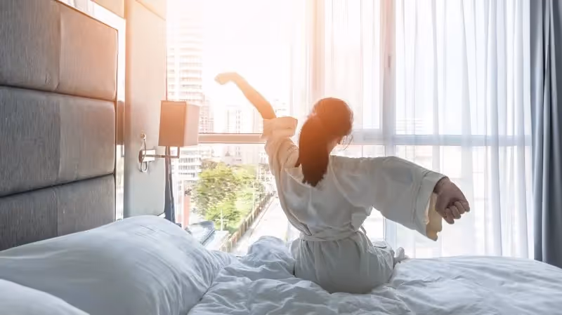 Femme en peignoir s’étirant au réveil dans une chambre d’hôtel lumineuse avec vue sur la ville.