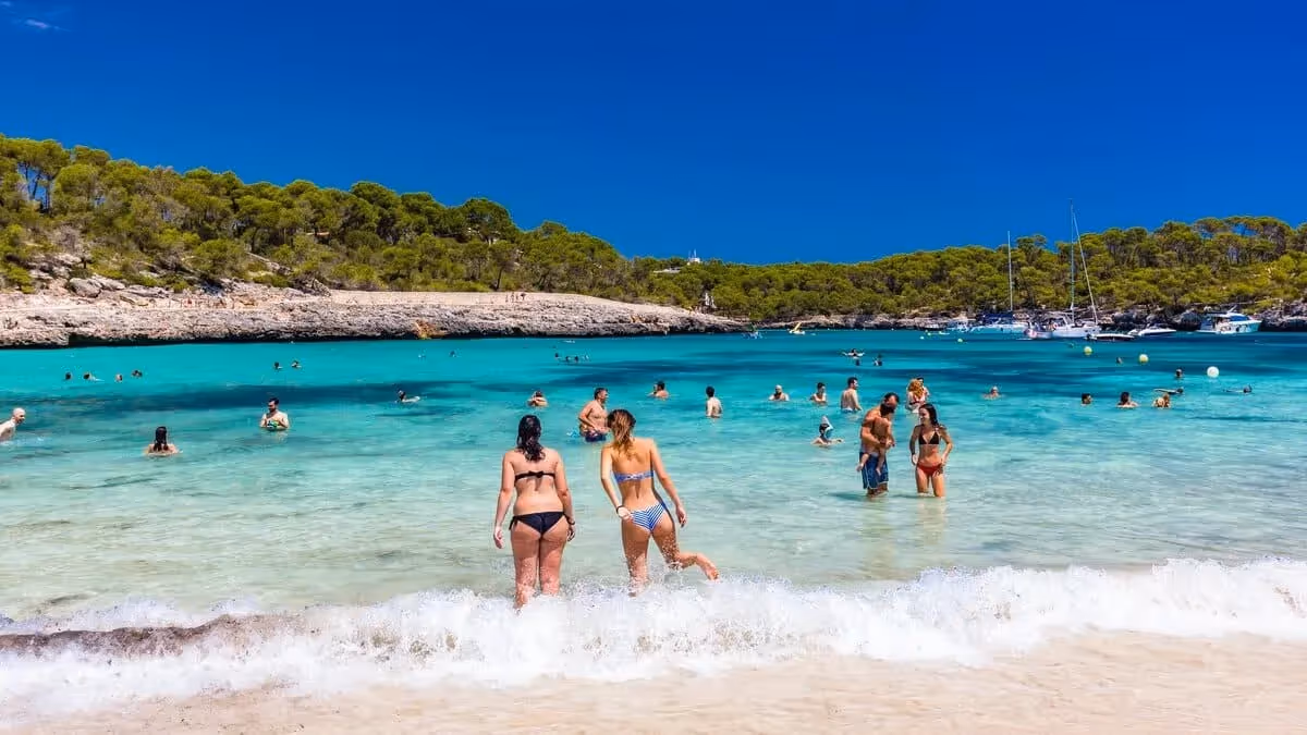 Vacanciers se baignant dans une crique aux eaux turquoise à Majorque, entourée de pins et de voiliers, sous un ciel bleu éclatant.