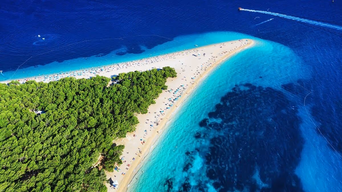 Vue aérienne de la plage Zlatni Rat à Bol, en Croatie, avec sa langue de galets entourée d’une mer turquoise et d’une forêt de pins.
