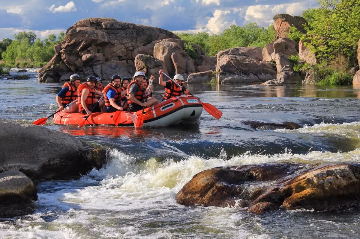 Groupe en gilets de sauvetage rouges faisant du rafting sur une rivière bordée de rochers, sous un ciel partiellement nuageux, pagaies rouges en main.
