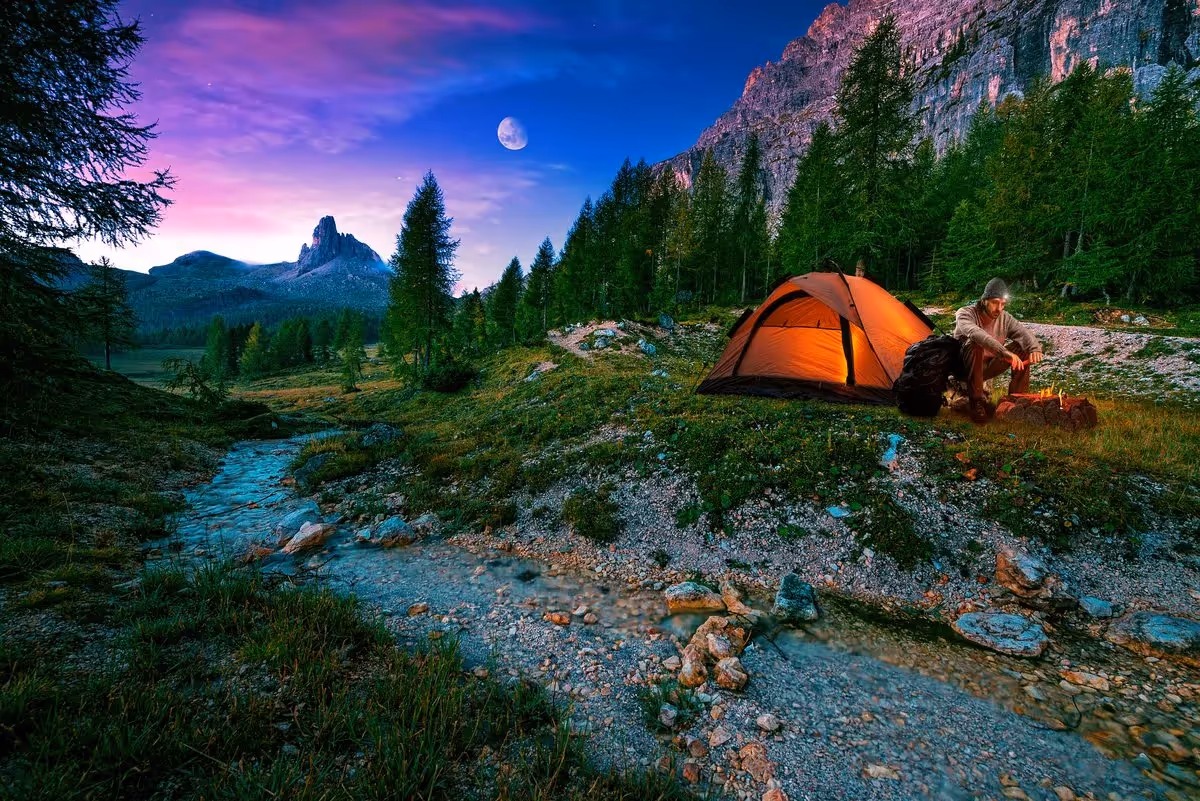 Homme en bivouac au bord d’un ruisseau en montagne, sous une tente éclairée au crépuscule