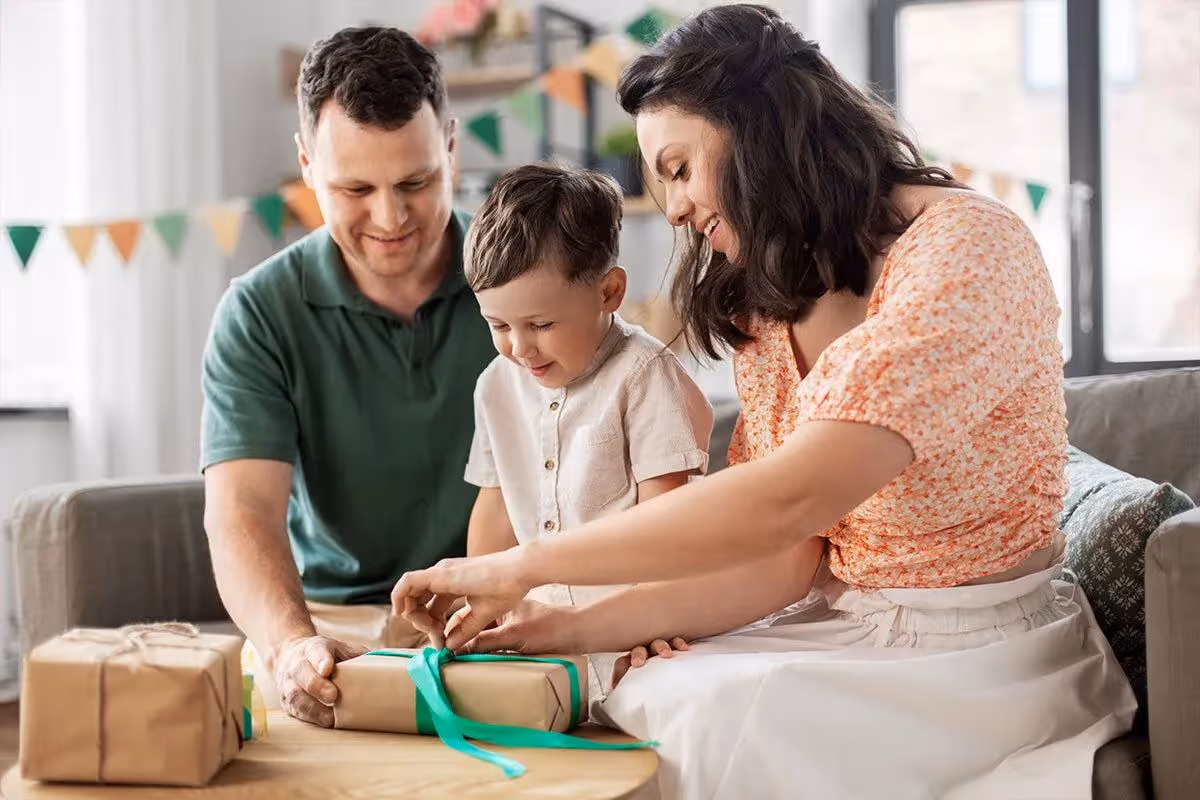 Un enfant ouvre un cadeau en famille, accompagné de ses parents souriants, dans un salon décoré — moment chaleureux avant un départ en voyage.