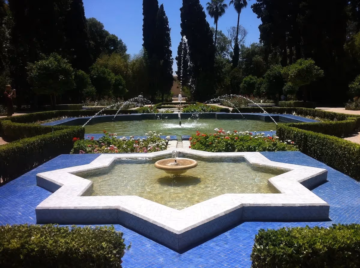 Fontaine décorative en étoile au cœur d’un jardin andalou, entourée de haies fleuries et de jets d’eau symétriques sous un ciel bleu.