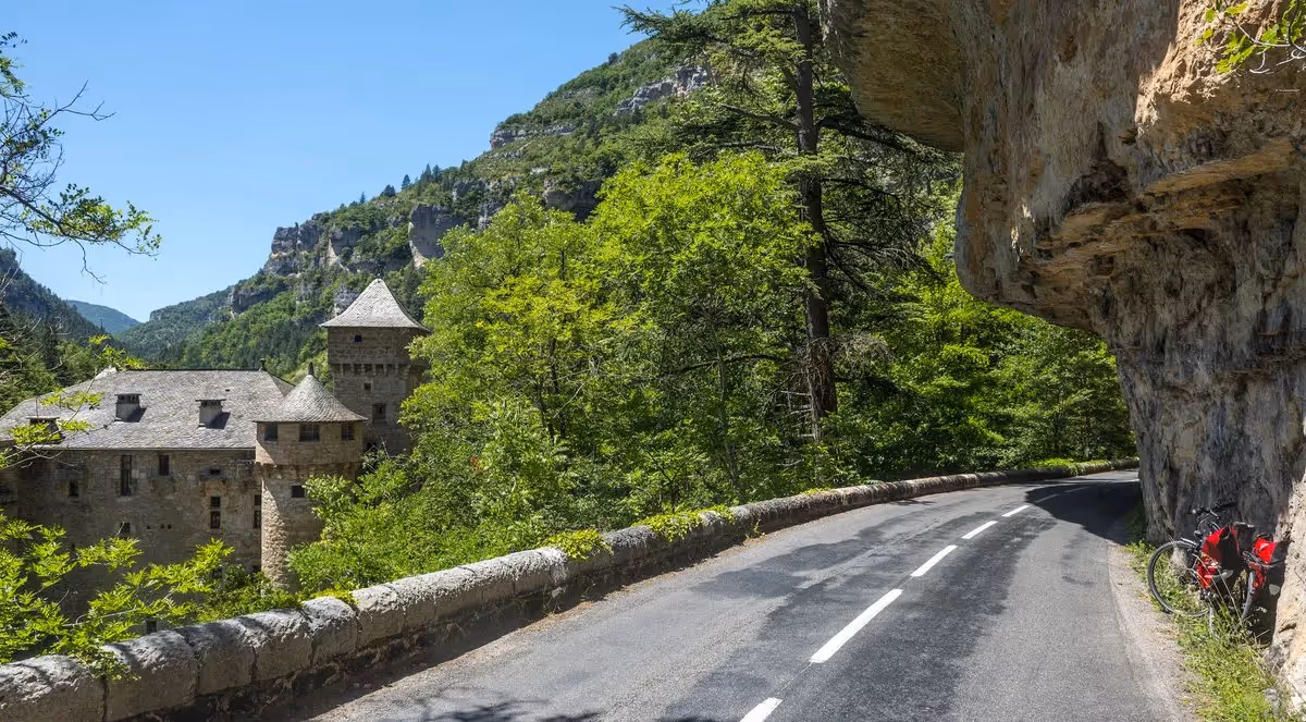 Route sinueuse bordée de falaises et forêt, avec un château en pierre niché dans les gorges des Cévennes.