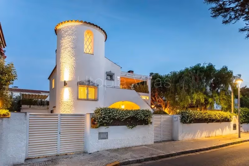 Façade élégante d'une villa blanche de style méditerranéen à Sitges, avec tour arrondie et éclairage doux en soirée.
