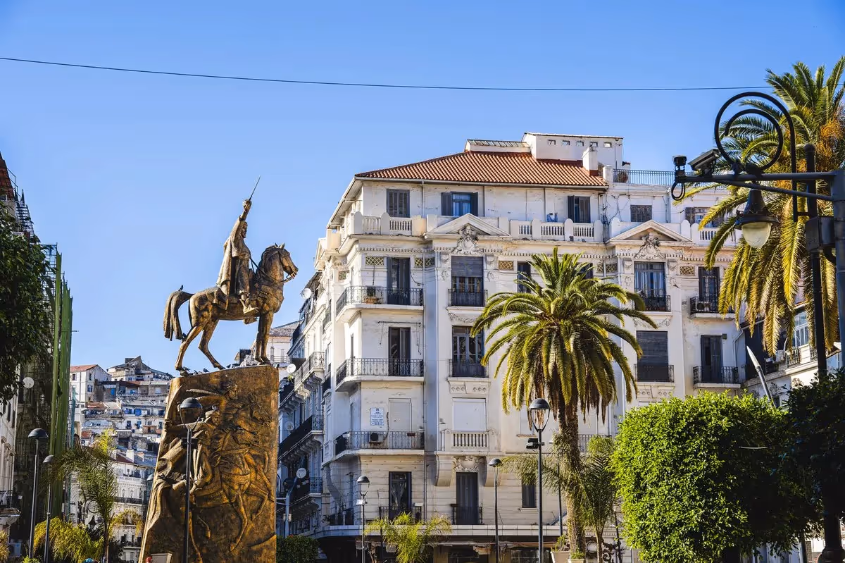 Statue de l’émir Abdelkader à cheval au centre-ville d’Alger, entourée d’immeubles haussmanniens et de palmiers sous un ciel bleu clair.