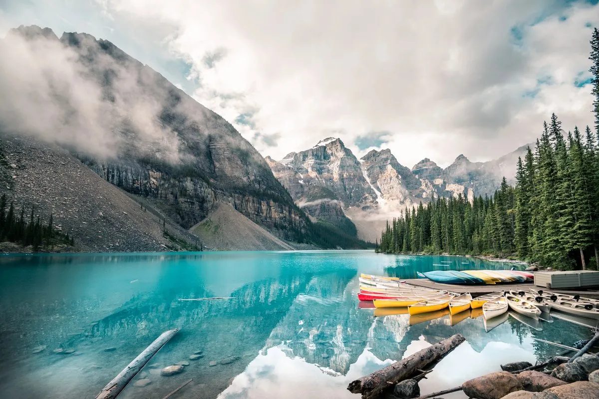 Vue sur le lac Moraine aux eaux turquoise entouré de sapins et des sommets enneigés des Rocheuses canadiennes, avec des canoës colorés au premier plan.