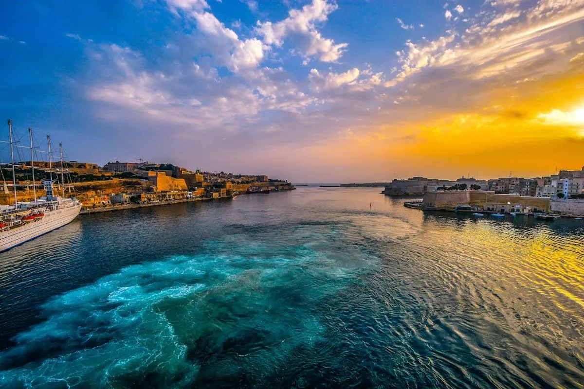 Vue panoramique sur un port fortifié de Malte au coucher du soleil, avec des eaux turquoise et un ciel aux teintes dorées.