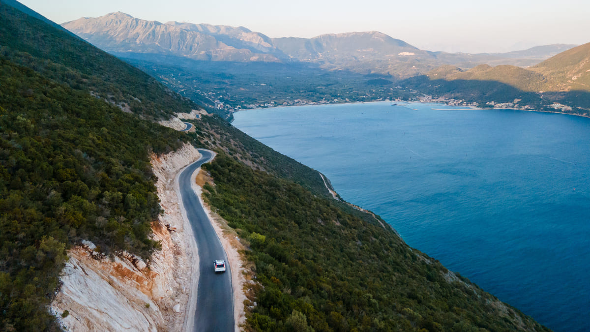 Vue aérienne d’une route côtière sinueuse en Épire, au nord-ouest de la Grèce, longeant une montagne verdoyante avec la mer Ionienne en contrebas, parfaite pour une micro-aventure estivale loin des foules.