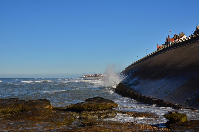 Vagues frappant la digue de Wimereux sur la Côte d’Opale, avec des maisons en arrière-plan sous un ciel dégagé.