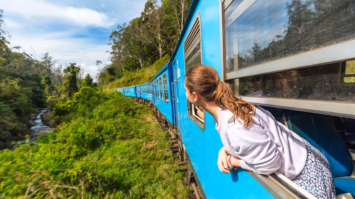 Voyageuse regardant le paysage depuis un train bleu traversant une région verdoyante, symbole du slow travel immersif.