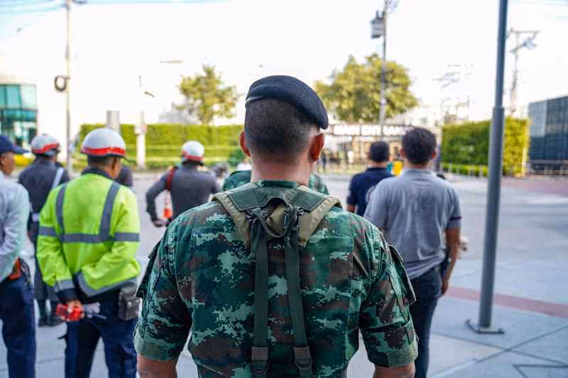 Soldat en uniforme camouflé vu de dos, entouré de travailleurs civils portant des casques de sécurité, dans un environnement urbain.