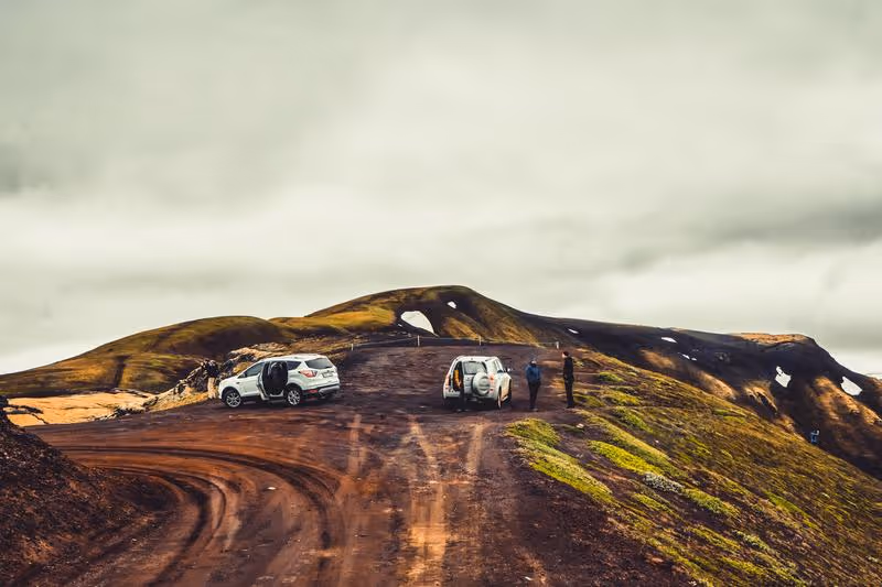 Véhicules tout-terrain arrêtés sur une piste de montagne volcanique en Islande, sous un ciel nuageux, avec des randonneurs observant le paysage.