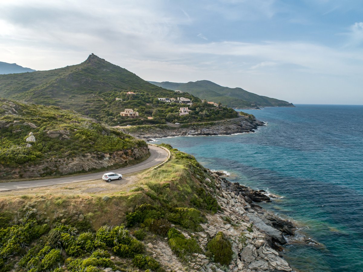 Vue aérienne d'une route côtière sinueuse en Corse longeant la mer Méditerranée, avec une voiture blanche, des collines verdoyantes et des maisons typiques nichées dans le maquis.