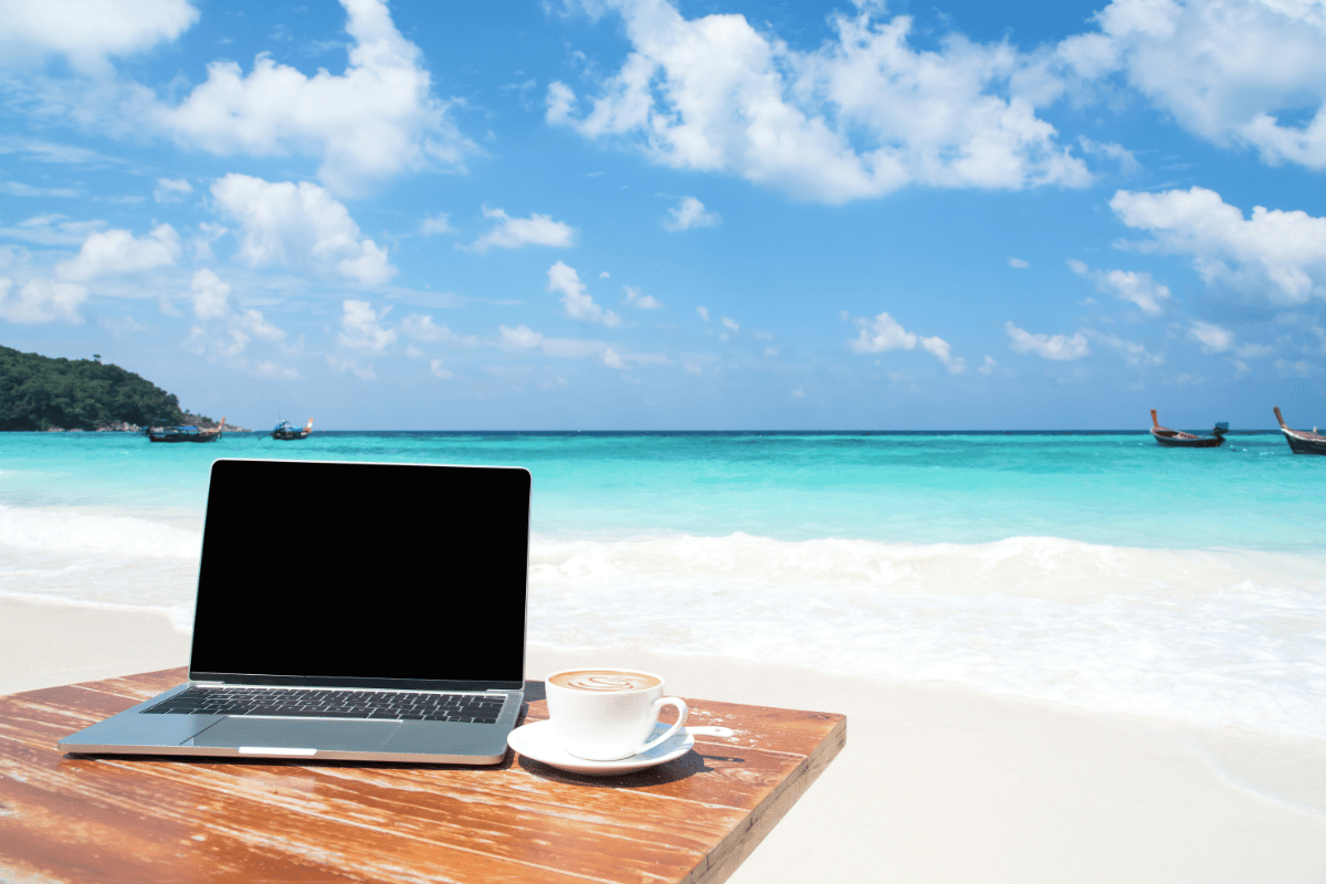 Ordinateur portable et tasse de café sur une table en bois, face à une plage de sable blanc et une mer turquoise sous un ciel bleu, illustrant le travail nomade en bord de mer.