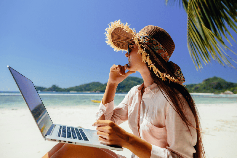 Femme portant un chapeau de paille travaillant sur un ordinateur portable, assise sur une plage de sable blanc avec vue sur la mer turquoise et les collines verdoyantes, illustrant le mode de vie nomade digital. 