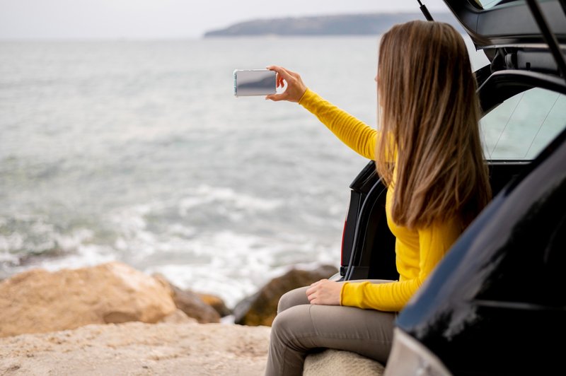 Femme prenant un selfie au bord de la mer lors d’un road trip, assise à l’arrière de sa voiture.
