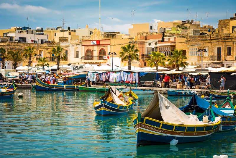 Port de pêche à Malte avec les traditionnels bateaux colorés luzzu et marché animé en arrière-plan.