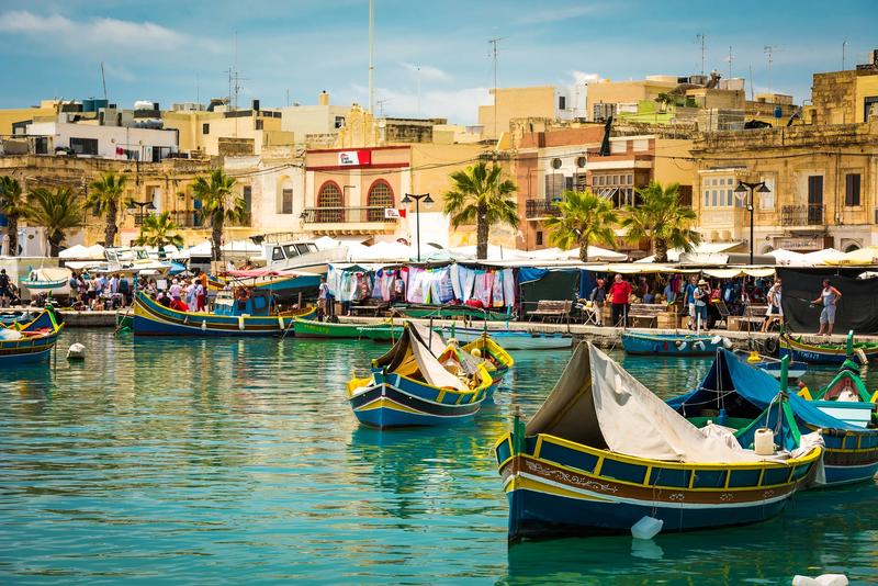 Port de pêche à Malte avec les traditionnels bateaux colorés luzzu et marché animé en arrière-plan.