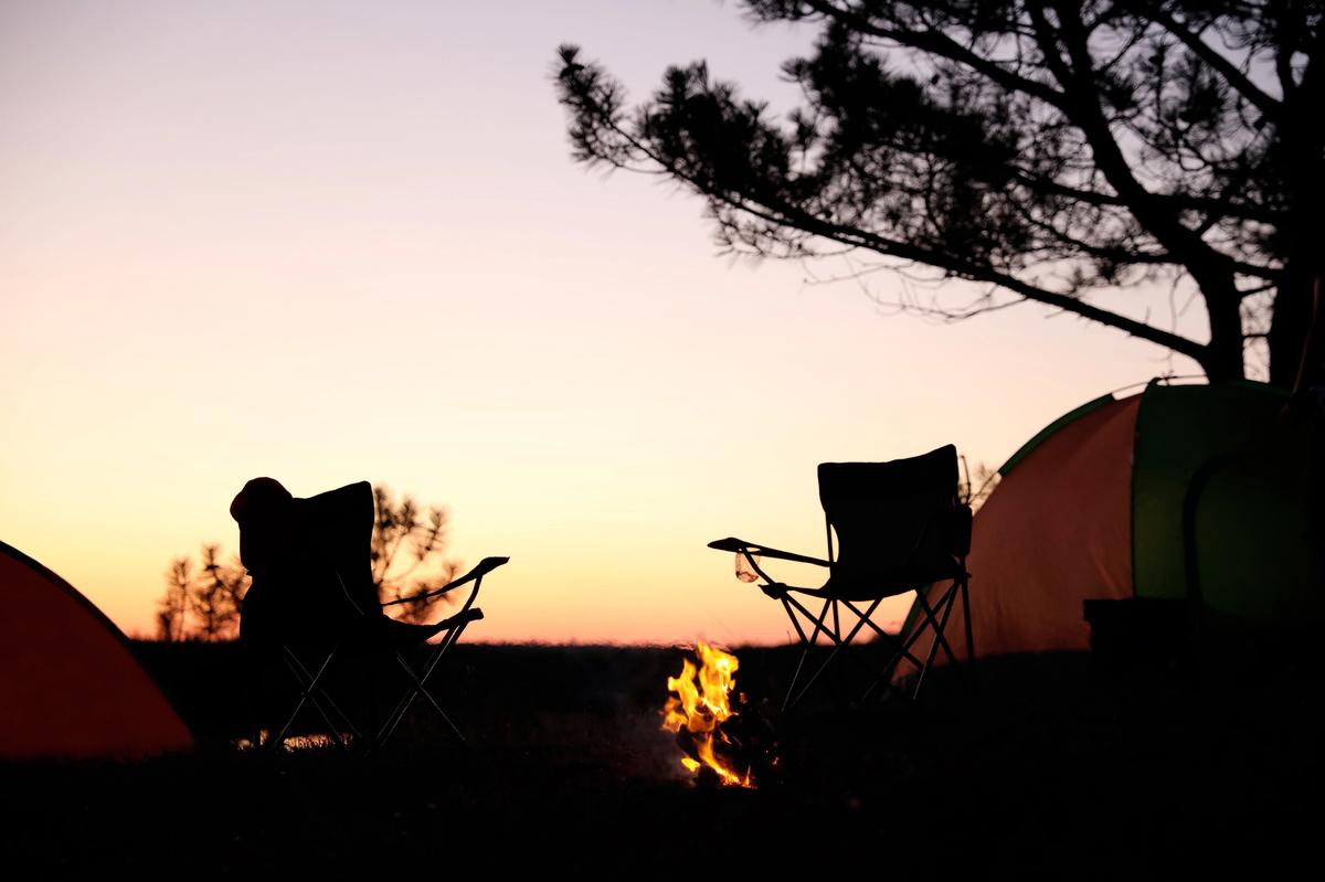 Campement au coucher du soleil avec deux chaises de camping et un feu allumé devant une tente.
