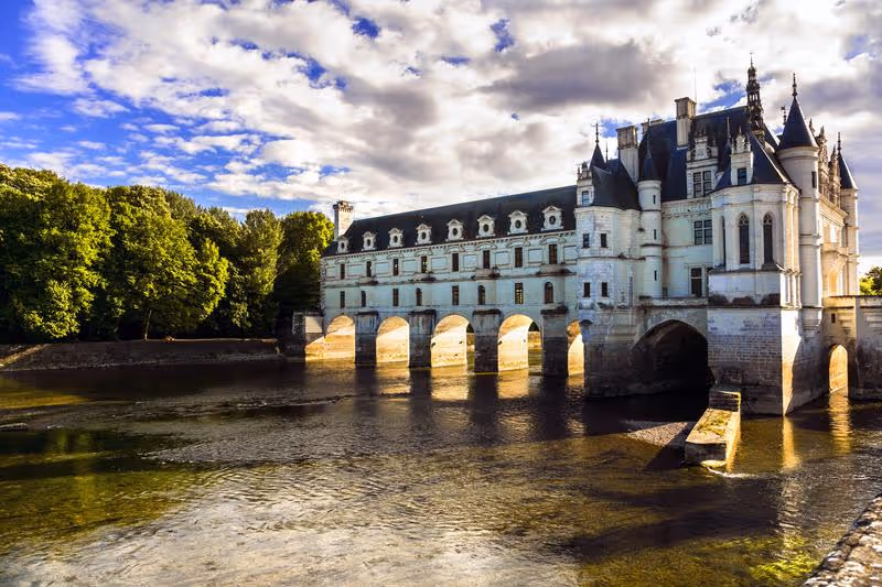 Château de Chenonceau en bord de rivière avec ses arches reflétées dans l’eau et entouré de verdure.