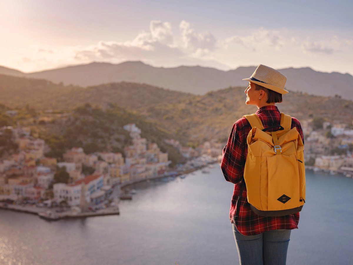 Voyageuse avec un sac à dos jaune contemplant un village méditerranéen en bord de mer au coucher du soleil.
