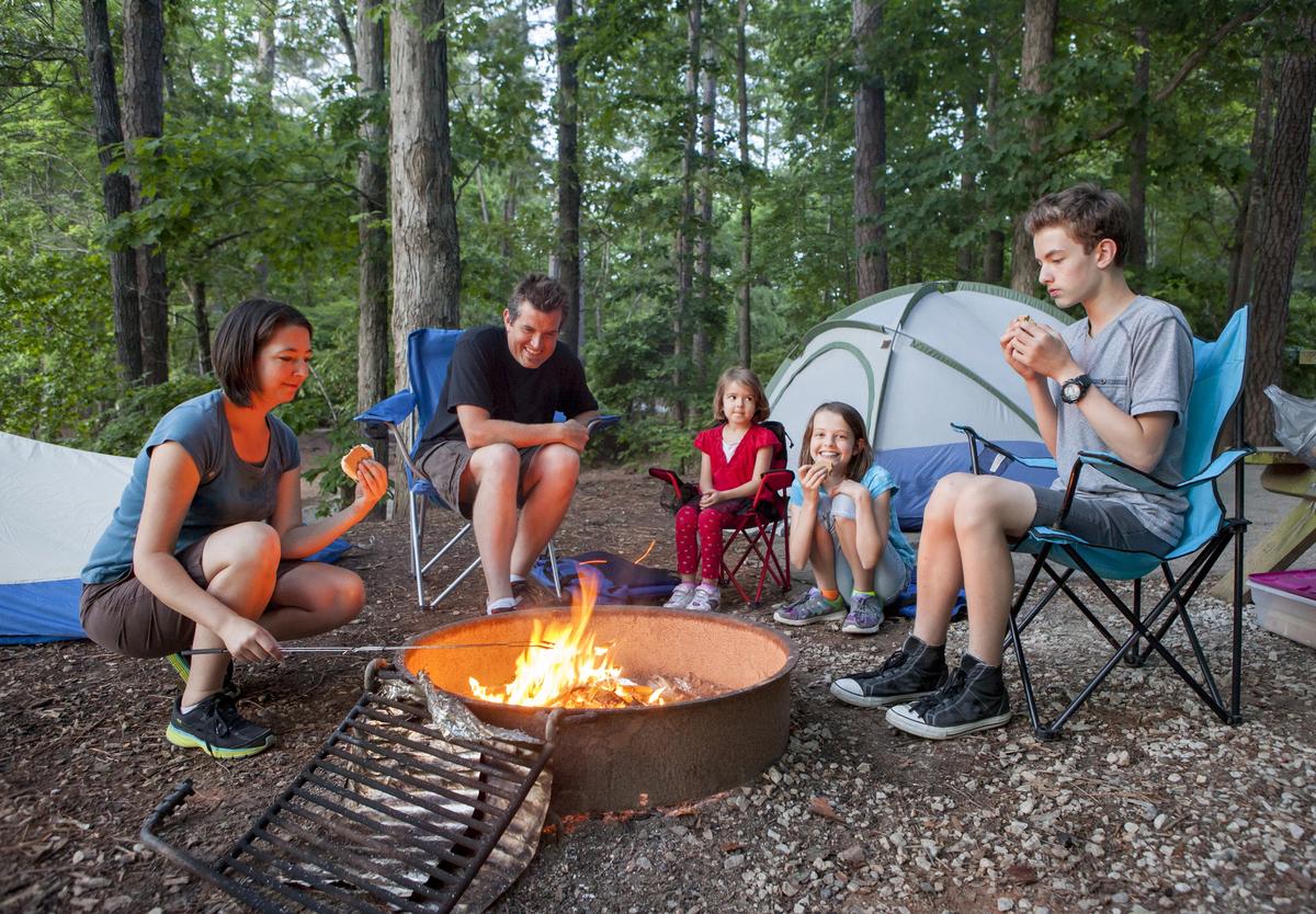 Famille en camping assise autour d’un feu de camp, préparant et dégustant des marshmallows devant une tente en forêt.