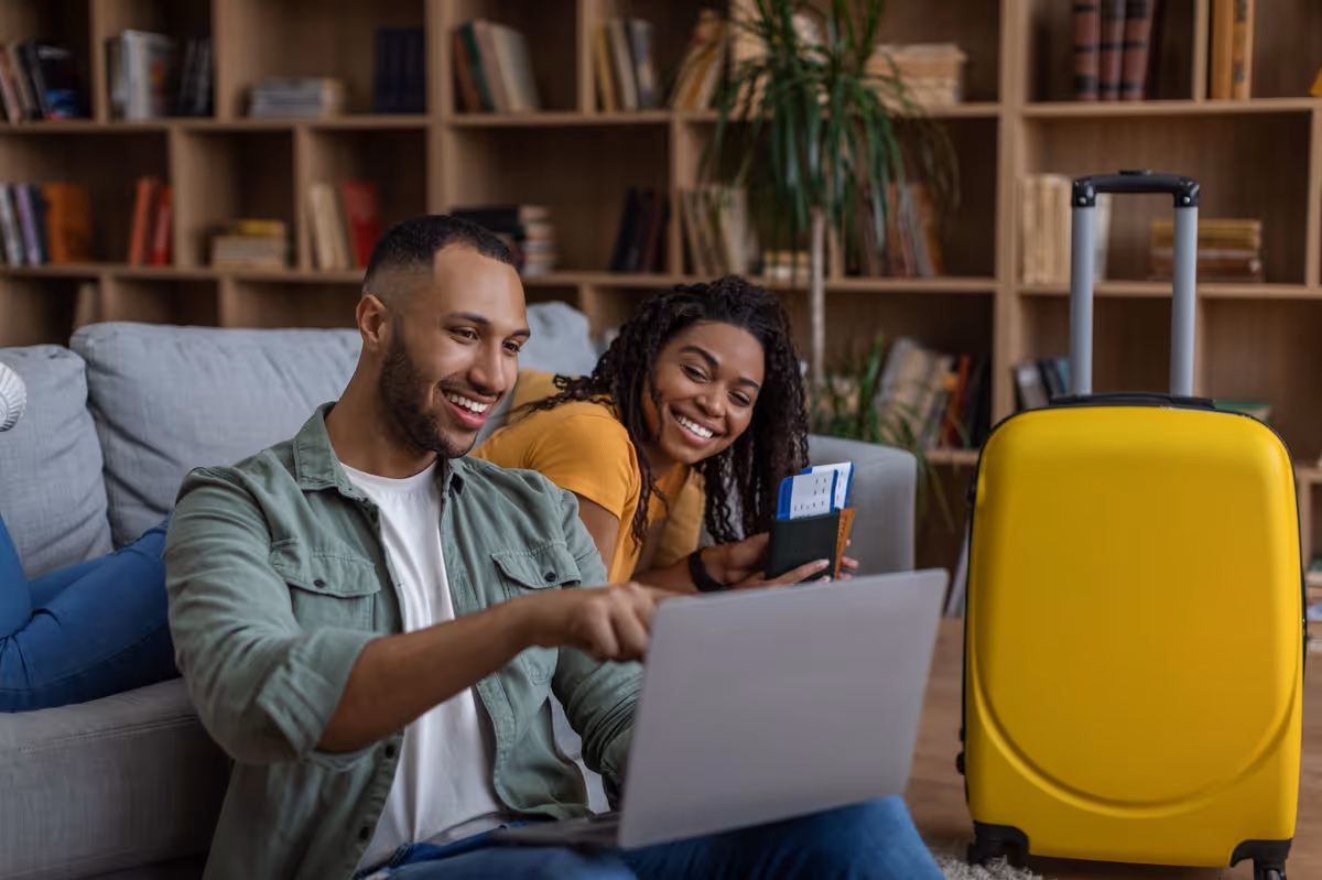 Un couple souriant assis sur un canapé prépare son voyage avec un ordinateur portable, une valise jaune et des billets d’avion.