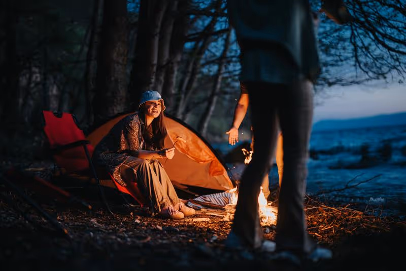 Scène de camping en bord de lac avec feu de camp et tente, moment convivial typique d’un road trip.